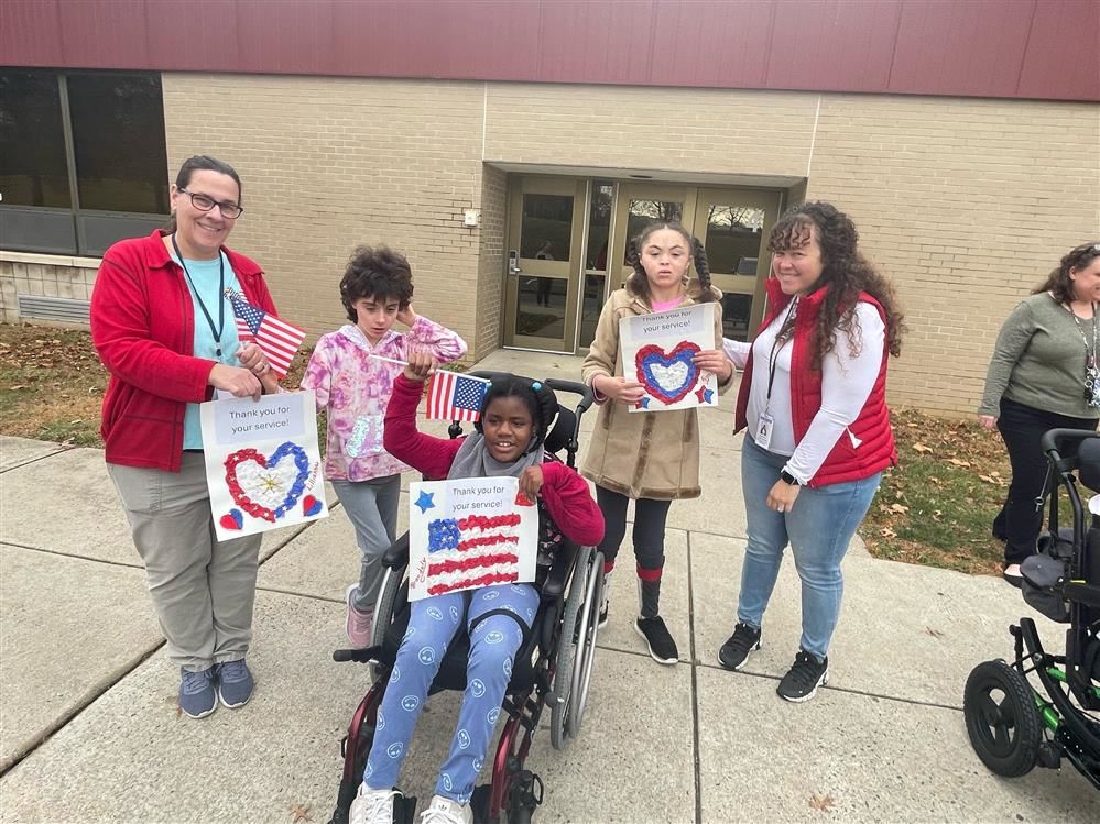  Two teachers are with three students as they hold up signs celebrating Veteran's Day.